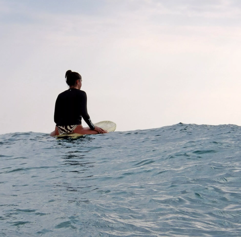 A woman is sitting on a surfboard in the ocean, facing away from the viewer. She is wearing a black long-sleeved shirt and patterned bottoms. The water is calm with small waves, and the sky is overcast. The image is shot from a low angle, emphasizing the vastness of the ocean and the woman's connection to it.